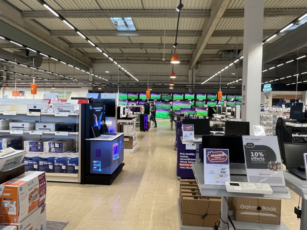 Großer Raum mit Elektronik einschließlich Computern, Druckern und Kartons in Regalen angeordnet, Menschen auf dem Boden, eine Säule, Schilder, Deckenlampen und Deckenventilatoren in einem Walmart-Laden.