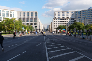 Eine belebte Straßenkreuzung in Berlin, Deutschland, mit Fußgängern und Radfahrern auf der Straße, hohen Gebäuden mit Fenstern, Bäumen, Laternenmasten und Schildern, unter einem bewölkten Himmel.