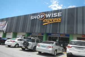 Ein Shopwise Express Geschäft mit parkenden Autos davor, mit einer Banner auf der linken Seite, unter einem bewölkten Himmel.