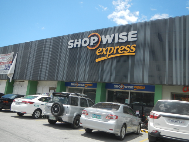 Ein Shopwise Express Geschäft mit parkenden Autos davor, mit einer Banner auf der linken Seite, unter einem bewölkten Himmel.