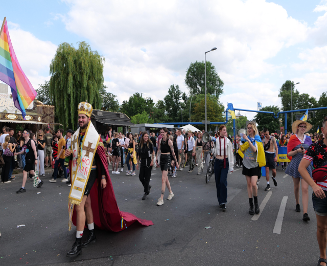 Eine Gruppe von Menschen marschiert bei der Pride Parade 2018 mit einer Regenbogenflagge und Musikinstrumenten, einige tragen Mützen, vor einem Hintergrund aus Laternenmasten, Bäumen, Hütten und einem bewölkten Himmel.