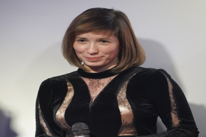 Frau in einem schwarzen Kleid, die ein Mikrofon hält, vor einer Wand im Hintergrund und einem selbstbewussten Gesichtsausdruck.