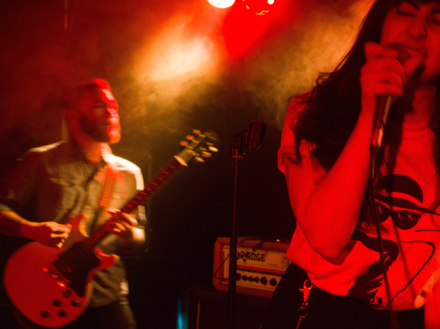 Eine Frau singt in ein Mikrofon auf der Bühne, während ein Mann Gitarre spielt und Deckenlampen das lebendige Setting erhellen.