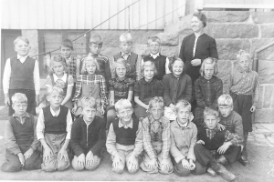 Schwarzes und weißes Foto einer Gruppe von Kindern, die vor einem Gebäude mit Treppe und Geländer posieren, mit dem Text "1930er Schulkinder" unten.