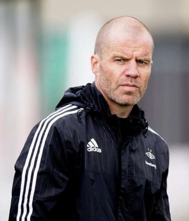 Ein kahlköpfiger Trainer in einer blauen Jacke mit weißen Streifen und weißem Kragen steht auf einem Fußballfeld, die Hände verschränkt, mit einem ernsten Gesichtsausdruck und einem unscharfen Hintergrund.