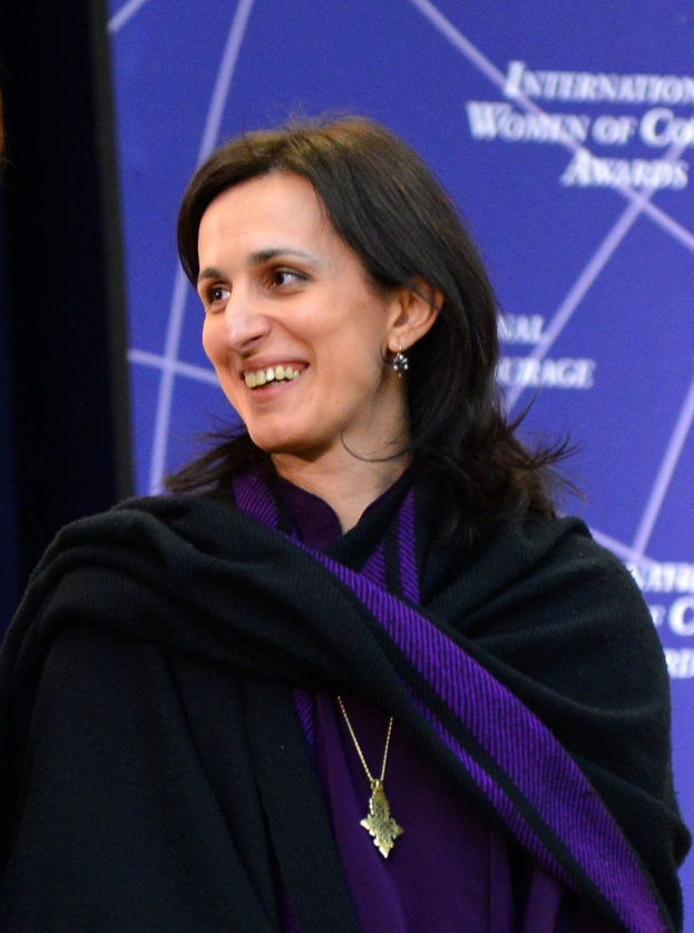 Eine Frau in einem lilafarbenen Kleid und einer schwarzen Stola steht neben einer anderen Frau, beide lächeln, mit einem Banner im Hintergrund, auf dem 'Internationale Frauen der Farbe Awards' steht.