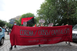 Eine Gruppe von Menschen marschiert mit einer roten Fahne, auf der "Corona-Krise/Kapitalismus" steht, eine Straße entlang. Parkende Fahrzeuge stehen auf beiden Seiten und im Hintergrund sind Gebäude, Bäume und ein klarer blauer Himmel zu sehen.