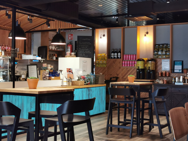 Innenraum eines Restaurants mit Holztischen, Stühlen, einer Topfpflanze auf einem Tisch, einer Schiefertafel, einem Kühlschrank, einem Feuerlöscher, Regalen mit Flaschen, Schränken mit Gegenständen, einem Rahmenfoto an der Wand, Deckenleuchten und Lampen.