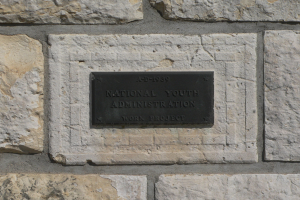 Steinwand mit einem rechteckigen Schild, auf dem "National Youth Administration" in weißer, fetter Schrift auf schwarzem Hintergrund steht.