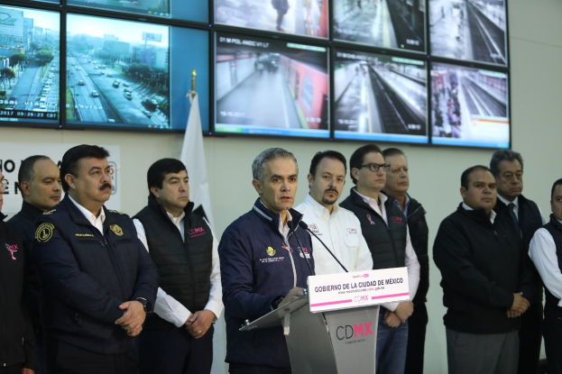 Gruppe von Männern, die vor einem Podium mit einem Mikrofon und einer Tafel mit der Aufschrift 'Mexikanische Zoll- und Grenzschutzbehörde' stehen, mit Fernsehern und einer Flagge im Hintergrund.