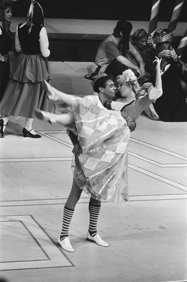 Schwarzes und weißes Foto von zwei Menschen, die auf einer Bühne tanzen, mit einem Pfahl im Vordergrund und einigen sitzenden Zuschauern auf einer Treppe im Hintergrund.