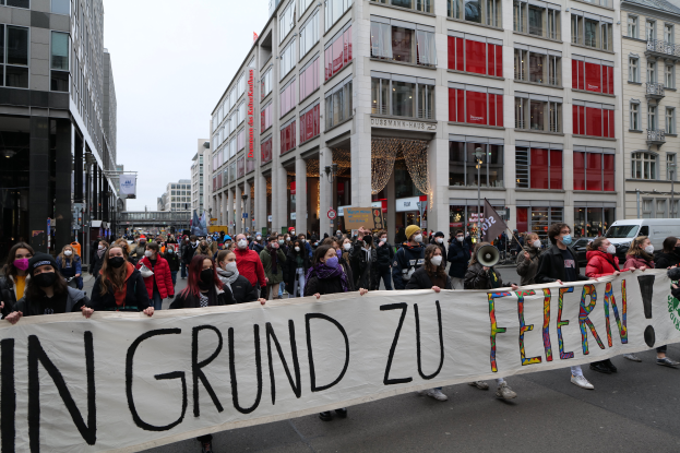 Eine Gruppe maskierter Personen hält ein Banner mit der Aufschrift "In Grund zu Feiern" vor Gebäuden, Laternenmasten und Fahrzeugen, was auf eine Protestaktion in Berlin, Deutschland, hindeutet.
