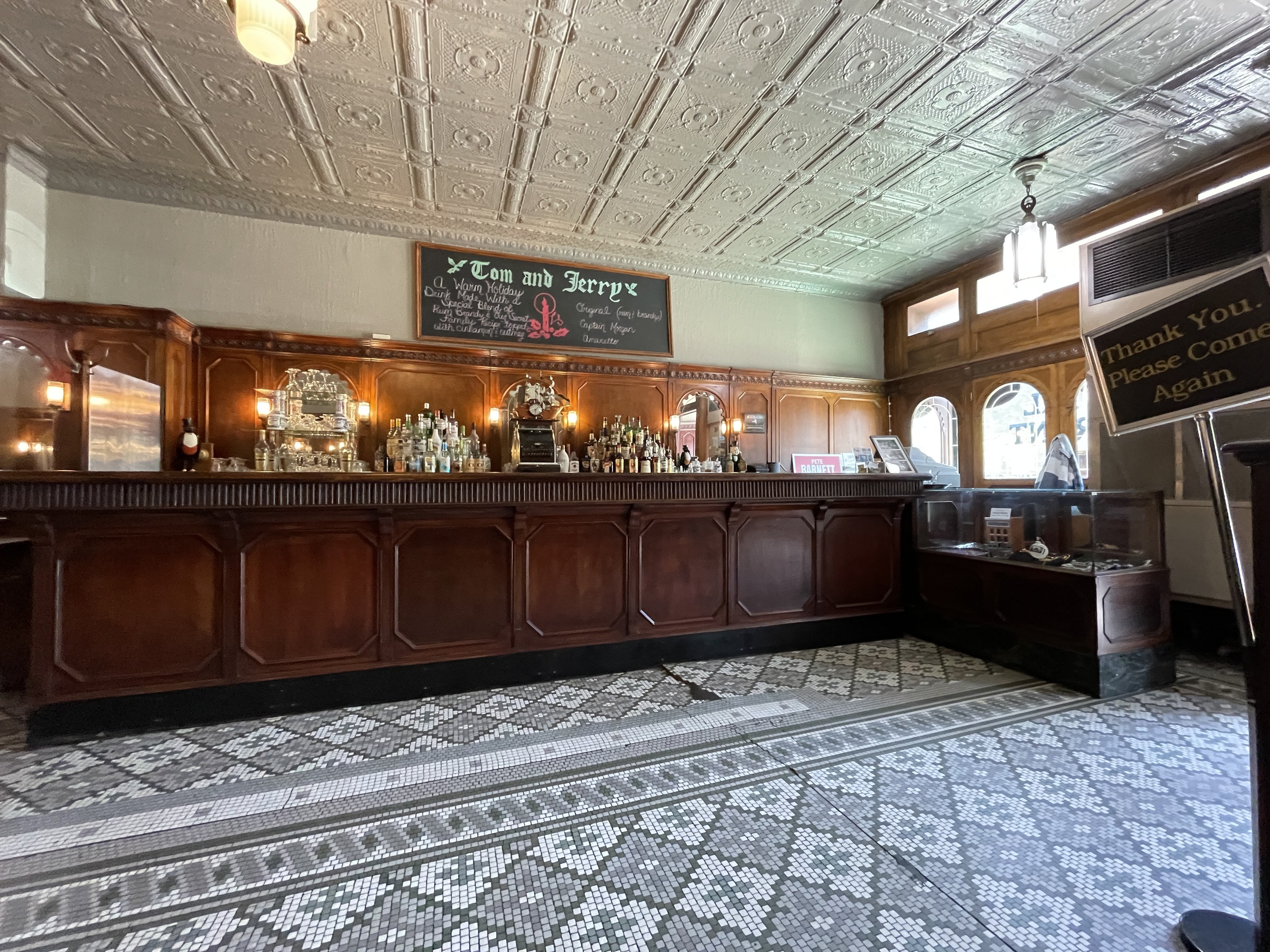 Ein großer Raum mit einer zentralen Bar, beleuchtet von Deckenlampen, mit Flaschen auf einem Tisch davor und einer Tafel mit Text auf der rechten Seite, im Tom and Jerry Pub in London.