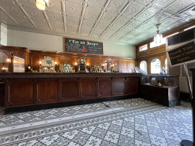 Ein großer Raum mit einer zentralen Bar, beleuchtet von Deckenlampen, mit Flaschen auf einem Tisch davor und einer Tafel mit Text auf der rechten Seite, im Tom and Jerry Pub in London.
