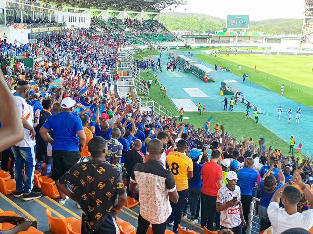 Eine große Menschenmenge versammelt sich vor einem eingezäunten Fußballfeld unter einem klaren blauen Himmel mit Hügeln im Hintergrund und wartet auf ein Spiel während der Afrikameisterschaft 2019.