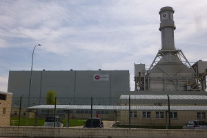 Ein großes Gebäude mit einem zentralen Turm, umgeben von einem Zaun, mit Fahrzeugen auf der benachbarten Straße, Masten, Lichtern, Drähten, Bäumen und Gras im Vordergrund und einem bewölkten Himmel im Hintergrund, das eine neue Energiespeicherkapazität repräsentiert.