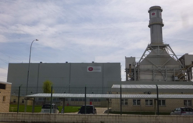 Ein großes Gebäude mit einem zentralen Turm, umgeben von einem Zaun, mit Fahrzeugen auf der benachbarten Straße, Masten, Lichtern, Drähten, Bäumen und Gras im Vordergrund und einem bewölkten Himmel im Hintergrund, das eine neue Energiespeicherkapazität repräsentiert.