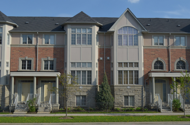 Eine Reihe von Stadthäusern in einem Wohnviertel mit sichtbaren Fenstern, Türen, Säulen, Geländern, Stufen, Pflanzen, Bäumen, Gras, einer Straße im Vordergrund und dem Himmel im Hintergrund.