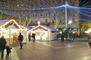 Ein lebendiger Weihnachtsmarkt in einer Stadt bei Nacht, mit Menschen, die herumlaufen, Ständen mit Lichtern und Gebäuden, Bäumen und Laternen im Hintergrund unter einem dunklen Himmel.