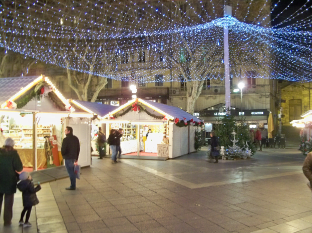 Ein lebendiger Weihnachtsmarkt in einer Stadt bei Nacht, mit Menschen, die herumlaufen, Ständen mit Lichtern und Gebäuden, Bäumen und Laternen im Hintergrund unter einem dunklen Himmel.