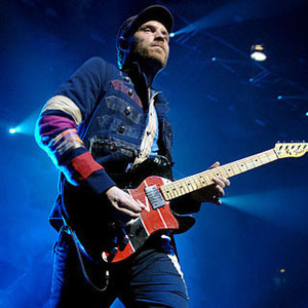 Ein Mann mit einer Mütze spielt Gitarre auf der Bühne eines Konzerts, beleuchtet von hellen Scheinwerfern, mit einem Ausdruck von Freude und Aufregung im Gesicht.