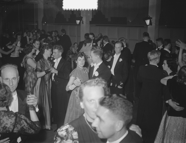 Eine Gruppe von Menschen, die auf einer Party in einem Schwarz-Weiß-Setting tanzen, mit Männern und Frauen verschiedenen Alters, einige halten Gläser, und eine beleuchtete Wand im Hintergrund.