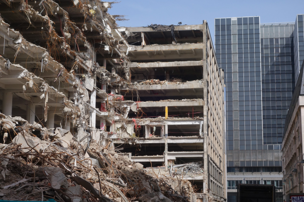 Ein große Abriss in der Mitte einer Stadt, umgeben von intakten Gebäuden, Straßeninfrastruktur, Fahrzeugen, Bäumen und einem klaren Himmel.