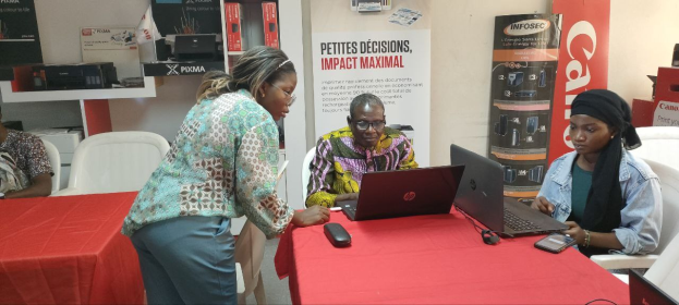 Eine Gruppe von Menschen sitzt um einen Tisch mit Laptops und Mobiltelefonen auf einer internationalen Geschäftsmesse in Senegal. Im Hintergrund sind Schilder, Schränke und eine Uhr zu sehen.
