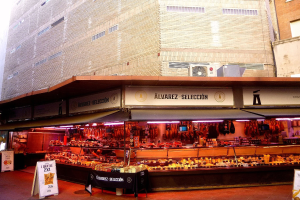 Ein belebter Markt mit verschiedenen Lebensmitteln, beleuchtet von Deckenlampen, umgeben von Texttafeln, mit Gebäuden und einem klaren blauen Himmel im Hintergrund.