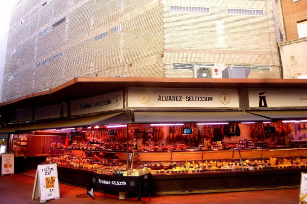 Ein belebter Markt mit verschiedenen Lebensmitteln, beleuchtet von Deckenlampen, umgeben von Texttafeln, mit Gebäuden und einem klaren blauen Himmel im Hintergrund.