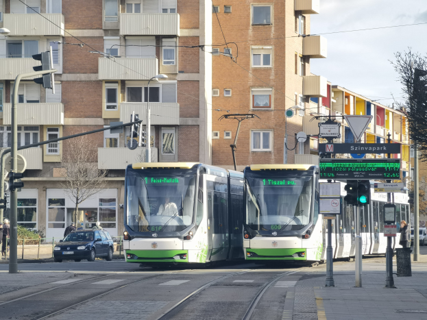 Zwei grüne und weiße Straßenbahnen fahren auf einer von hohen Gebäuden gesäumten Stadtstraße, umgeben von Fahrzeugen, Fußgängern, Bäumen und Straßeninfrastruktur unter einem bewölkten Himmel.