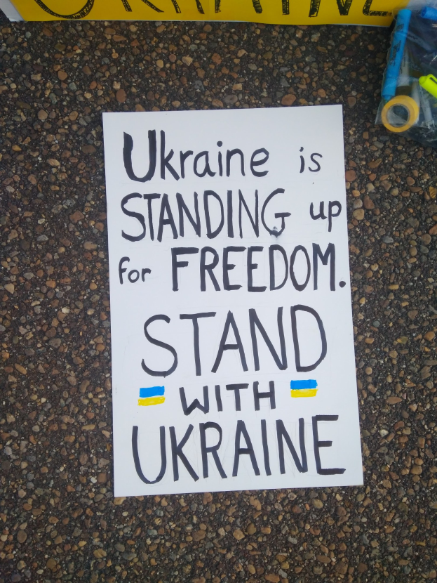 Weißer Zettel mit dem Text "Ukraine steht für die Freiheit ein, steht mit Ukraine" auf dem Boden vor einer gelben Fahne, rechts daneben ein Plastikbeutel mit verschiedenen Gegenständen.