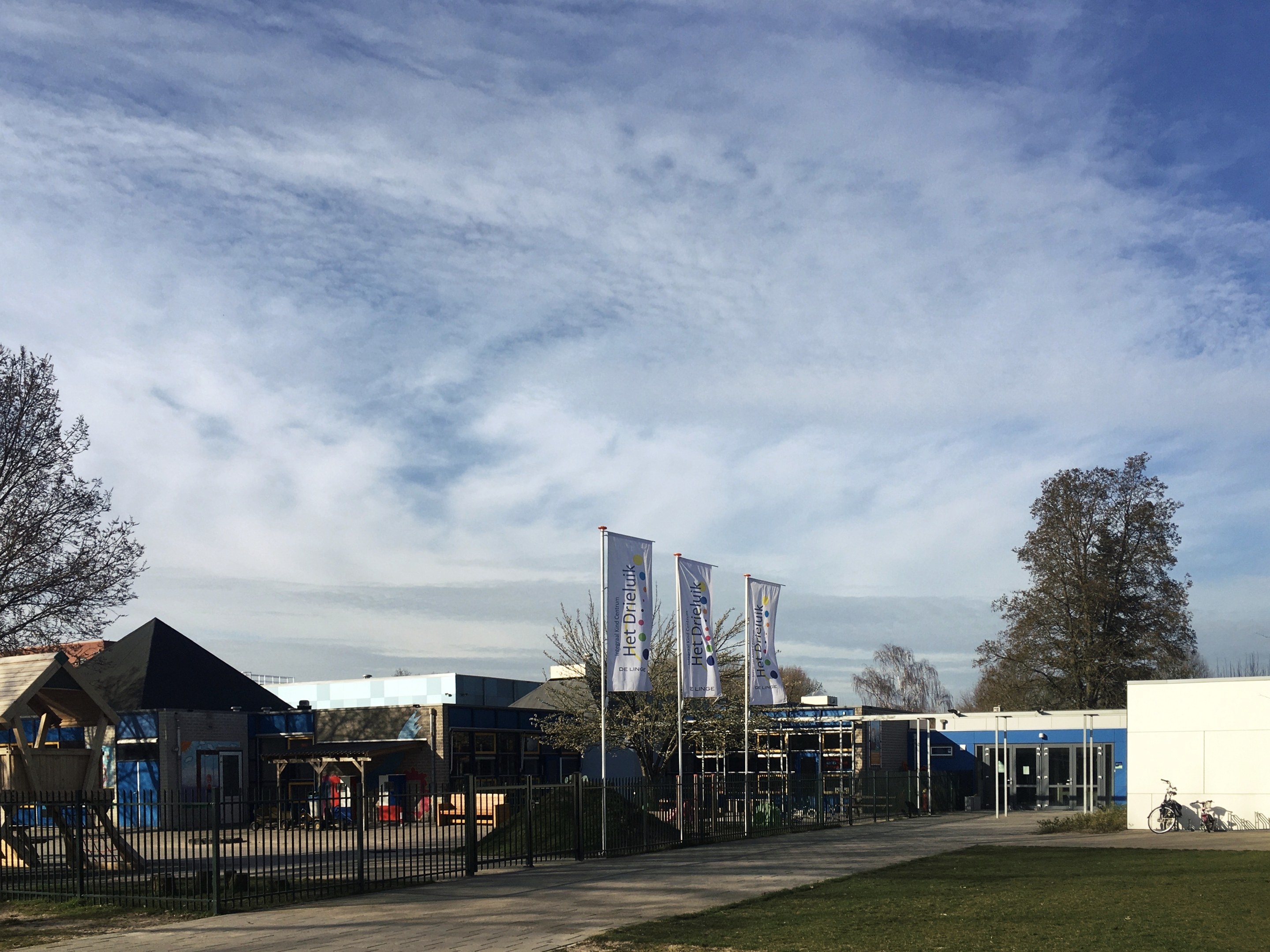Großes Gebäude mit einem grasigen Bereich davor, umgeben von einem Metallzaun, mit einem Weg, Bäumen, Pfosten mit Bannern, einem geparkten Fahrrad, nahen Häusern und einem bewölkten Himmel, als das neue Spielplatzgelände an der Schule identifiziert.