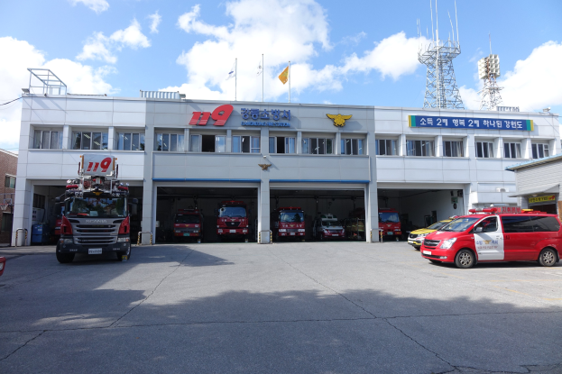 Feuerwache mit mehreren Feuerwehrautos vor dem Gebäude geparkt, Gebäude mit Text und Fenstern, Flaggen auf Stangen im Hintergrund, bewölkter Himmel und eine ferne Turm.
