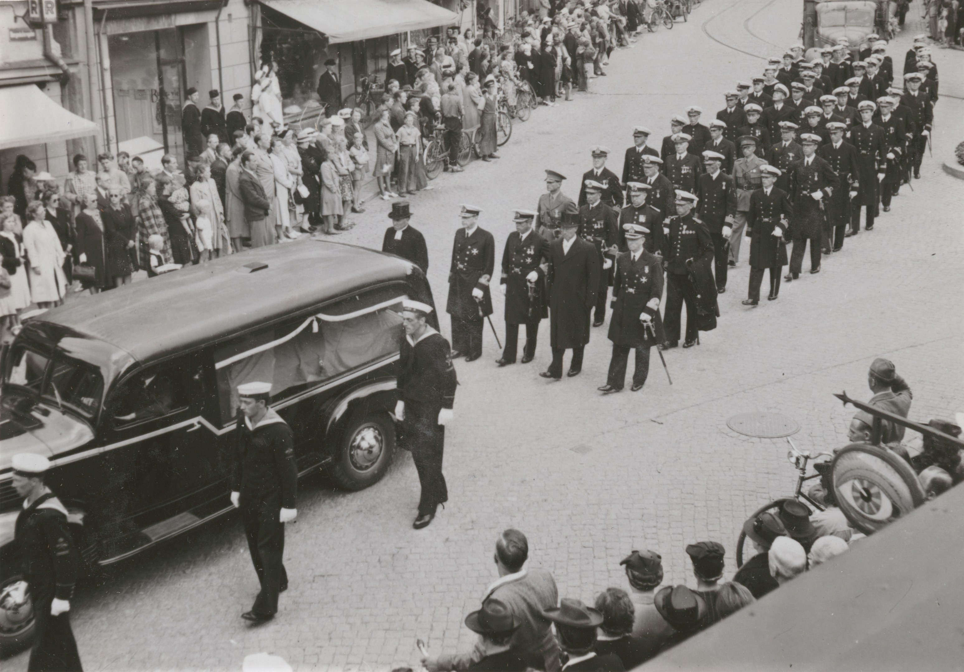 Schwarz-weißes Trauergeleit mit einem Leichenwagen, der einen Sarg trägt, umgeben von Menschen, einige halten Waffen, Fahrzeuge auf der Straße und Gebäude mit Fenstern und Namensschildern im Hintergrund.