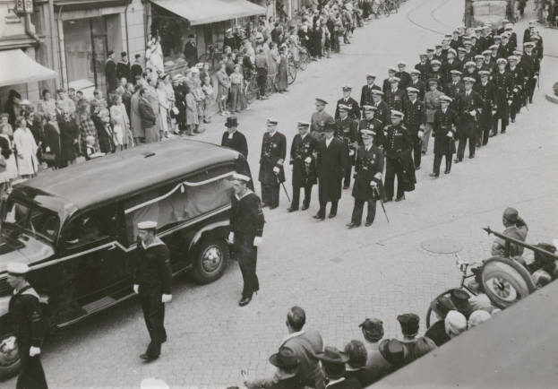 Schwarz-weißes Trauergeleit mit einem Leichenwagen, der einen Sarg trägt, umgeben von Menschen, einige halten Waffen, Fahrzeuge auf der Straße und Gebäude mit Fenstern und Namensschildern im Hintergrund.