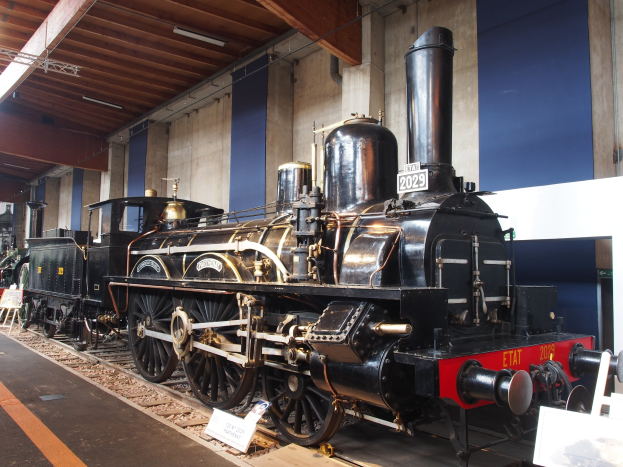 Alte Dampflokomotive auf Eisenbahnschienen in einem Museum ausgestellt, umgeben von Informationsschildern, St├Ąnden und anderen Ausstellungsstücken, mit einer beleuchteten Decke und Wand im Hintergrund.