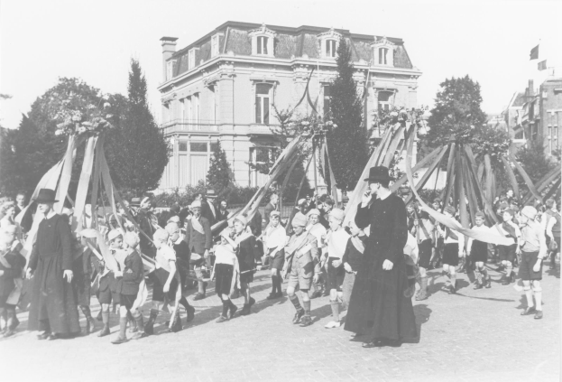 Ein Schwarz-Weiß-Bild einer Gruppe von Menschen, die vor einem Gebäude die Straße entlanggehen, einige halten Fahnen, tragen Hüte, mit Bäumen und Gebäuden im Hintergrund und einem sichtbaren Himmel, mit einem Maypole als Indikator für May-Day-Feierlichkeiten.