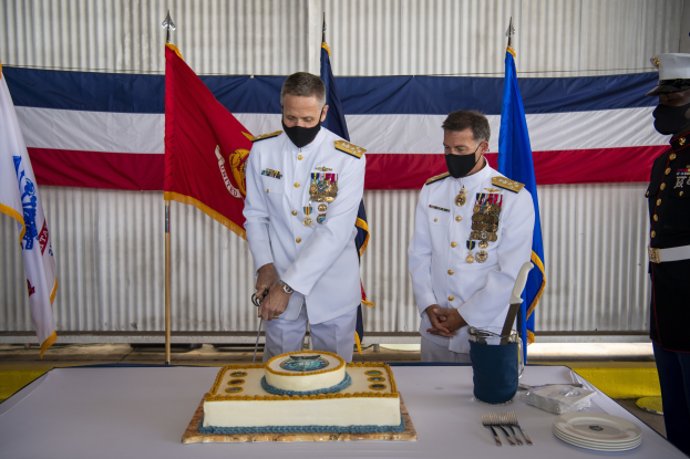 Zwei Männer in weißen Marineuniformen schneiden einen Kuchen bei einer 50-jährigen Feier, umgeben von Essbesteck und Fahnen.
