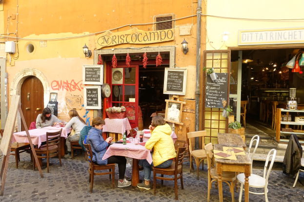 Eine Gruppe von Menschen sitzt an Tischen vor einem Restaurant in Rom, Italien, mit Flaschen, Gläsern und anderen Gegenständen auf den Tischen und Wänden mit Texttafeln, einer Tür, die ins Innere führt, und Beleuchtung im Bereich, mit Regalen mit verschiedenen Gegenständen im Inneren.