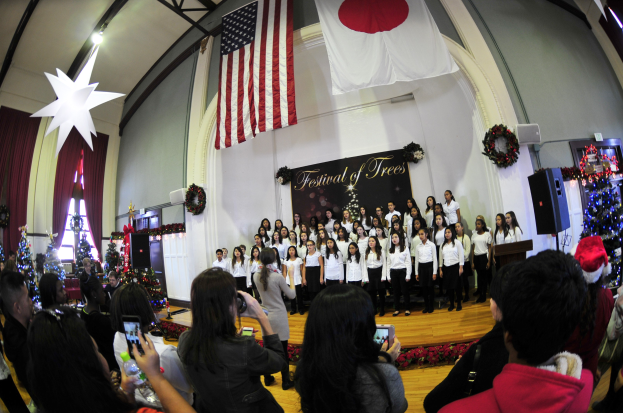 Eine Gruppe von Menschen auf einer Bühne mit Weihnachtsdekoration, einem Rednerpult, Lautsprechern, einer Texttafel und einem Publikum mit Kameras und Handys.