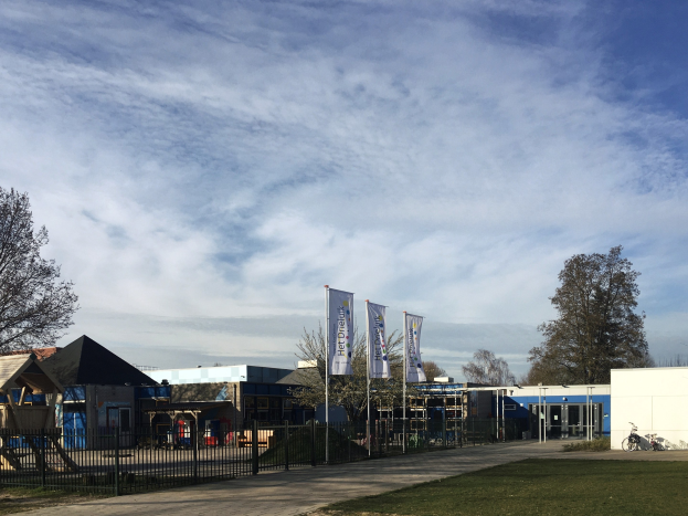 Großes Schulgebäude mit einem grasbewachsenen Bereich, der von einem Metallzaun umgeben ist, mit einem Weg, Bäumen, Bannern, einem geparkten Fahrrad, nahen Häusern und einem bewölkten Himmel.