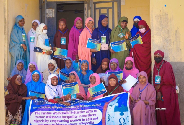 Gruppe von Frauen, die strahlend in die Kamera lächeln und Zertifikate in den Händen halten, vor einem Gebäude mit einer Tafel, auf der steht: "Funtua Women Wiki Initiative setzt sich für die Bekämpfung von Wikipedia-Ungleichheit im Norden Nigerias ein."