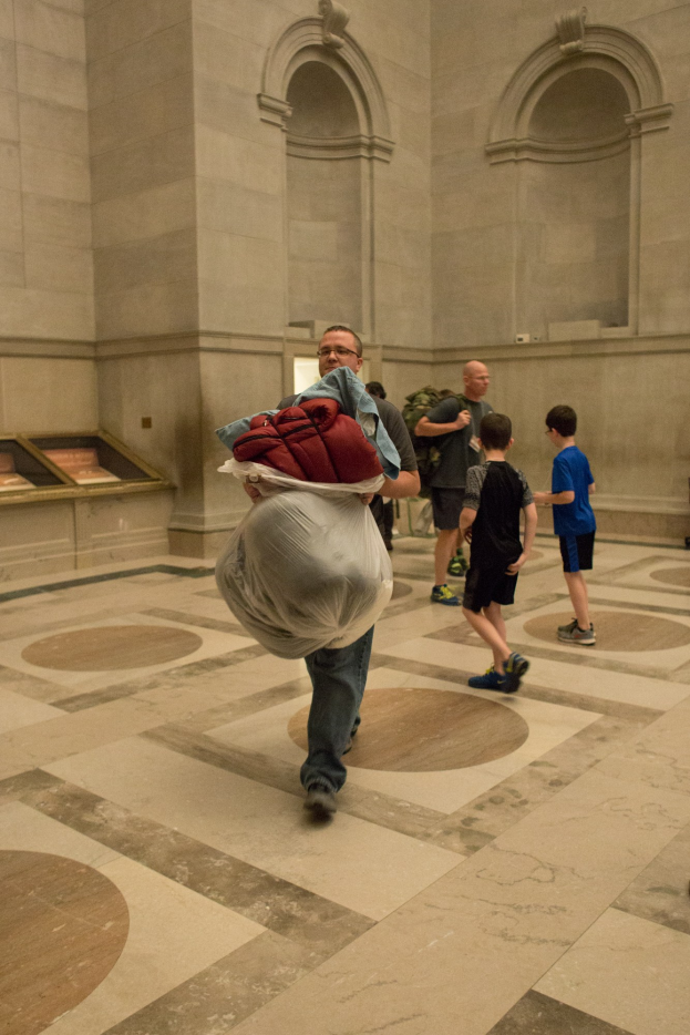 Ein Mann trägt eine Gepäcktasche in einem großen Raum mit einer Wand im Hintergrund, umgeben von ein paar stehenden Menschen, die Schuhe tragen.