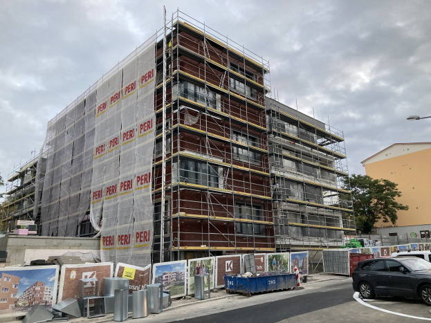 Großes Gebäude in Bau mit Gerüst, umgeben von Bäumen, Straßeninfrastruktur, Fahrzeugen und Werbetafeln vor einem bewölkten Himmel.