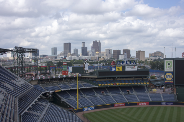 Baseballstadion mit einer Stadtkulisse im Hintergrund, gefüllt mit Stühlen, Pfosten, Tafeln mit Text und anderen Gegenständen, grasbewachsenem Boden, Bäumen und Gebäuden in der Ferne, bewölktem Himmel.