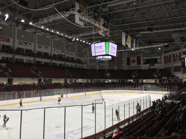 Ein Eishockeyspiel in einer großen Arena mit einem zentralen Netz, Zuschauern auf beiden Seiten und Banner und Flaggen an den Wänden, identifiziert als die St. Louis Blues Arena.