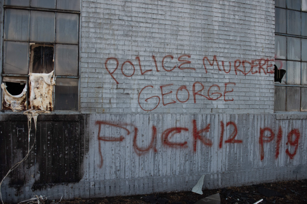 Graffiti auf einer Gebäudewand mit der Aufschrift "Polizei ermordet George Fuck 12 P19" mit Fenstern und verstreuten Gegenständen davor.