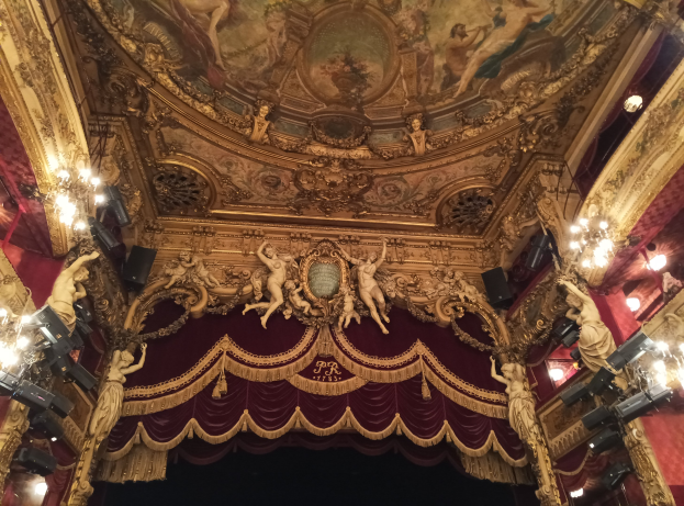 Theaterinnenraum mit roter Gardine, Kronleuchtern, Skulpturen, Lampen und einer Deckenmalerei, die eine Bühne mit einigen Personen am Boden zeigt.