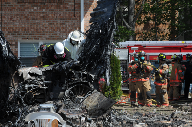 Feuerwehrleute in Schutzausrüstung löschen ein Gebäude Feuer, mit verkohlten Trümmern einschließlich eines Fahrzeugs, Stuhl, Pflanzen und Bäume sichtbar auf dem Boden.